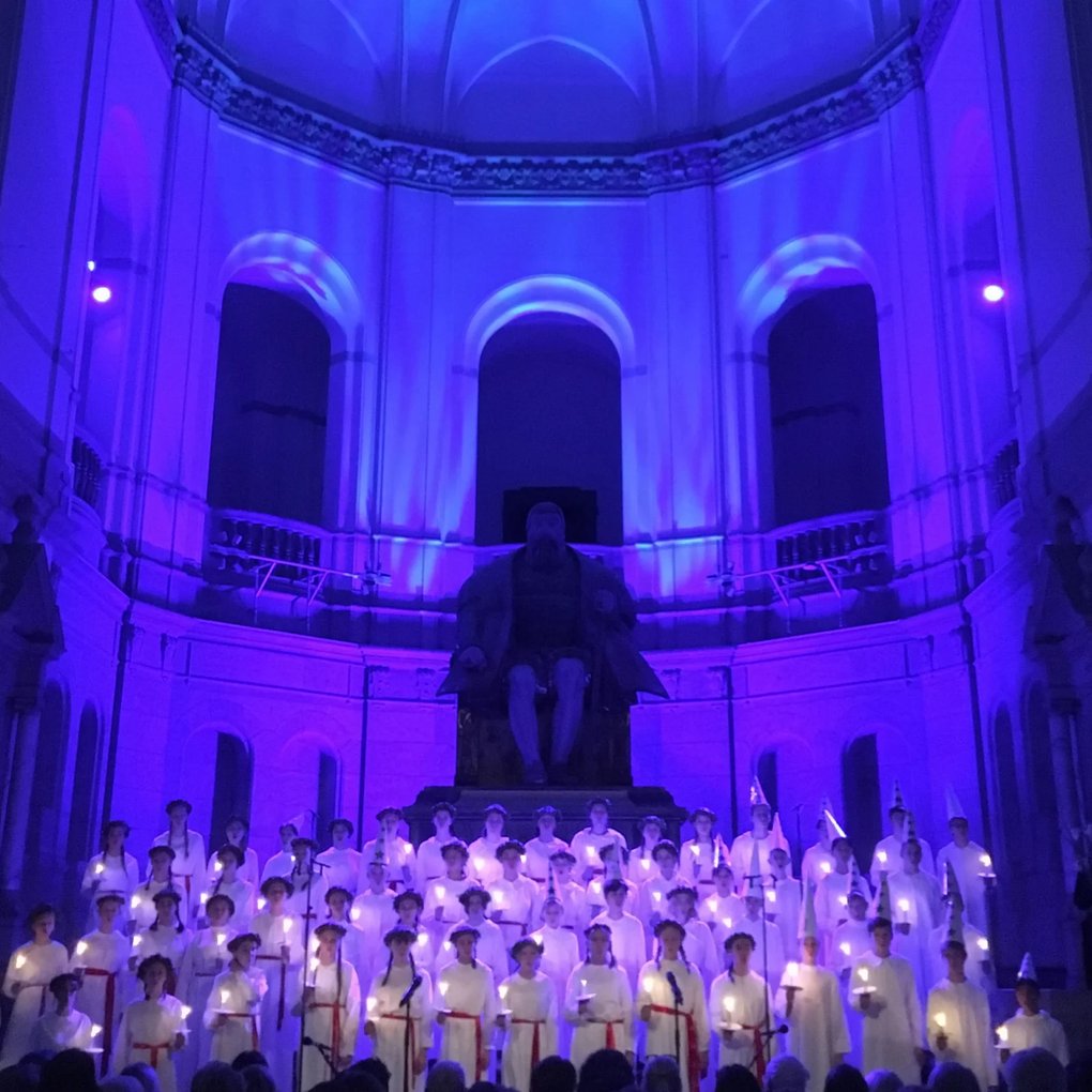 Traditional Lucia-choir in front of a large statue of Gustav Vasa in a great hall.